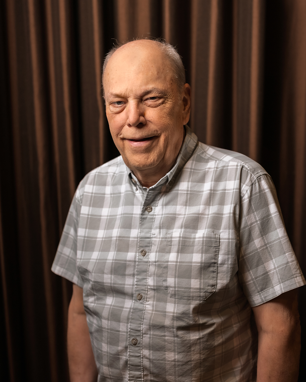 Elderly man smiling in plaid shirt indoors.