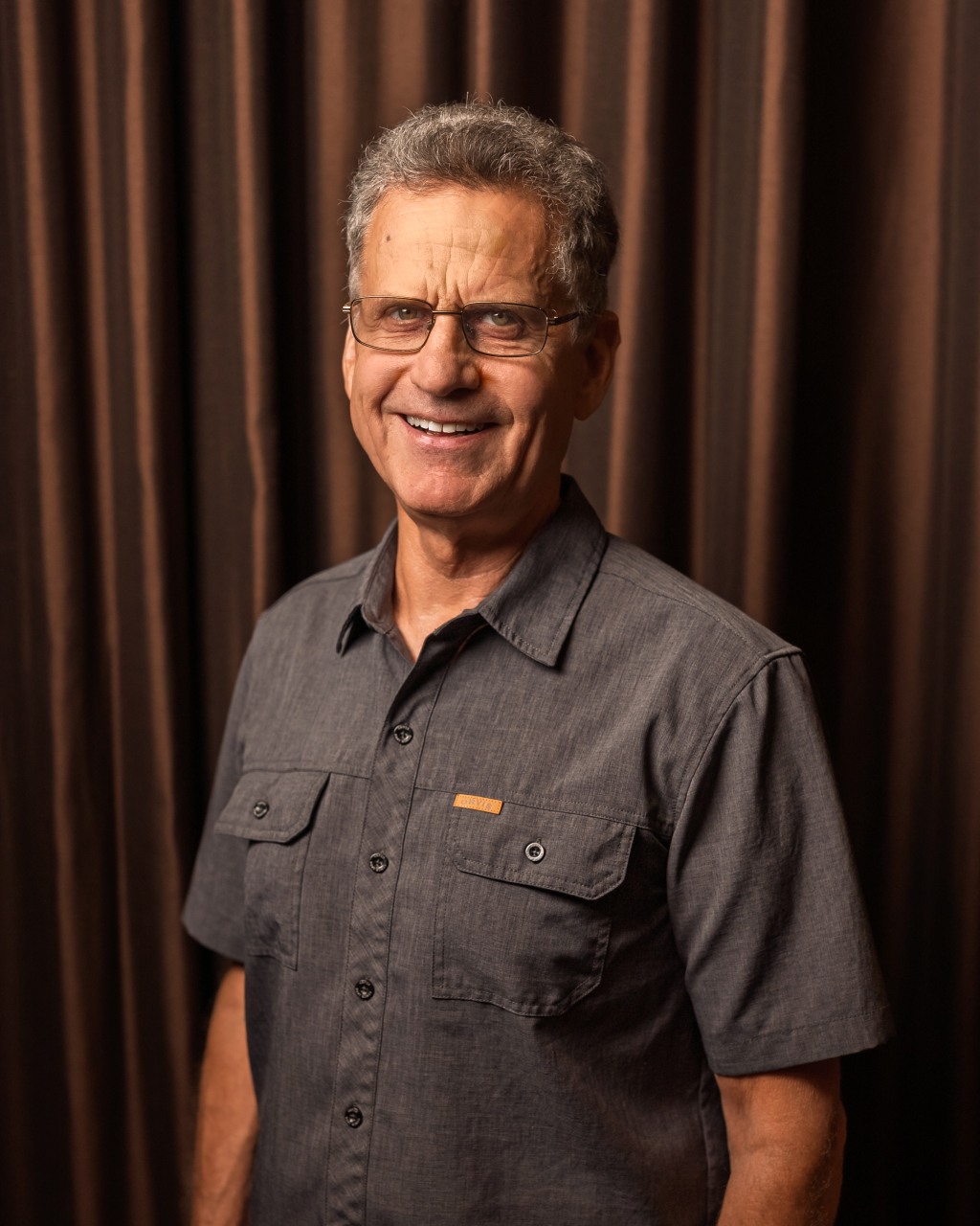 Smiling man in gray shirt, brown curtain backdrop.